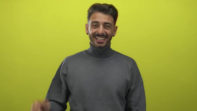 Young hispanic man wearing gray turtleneck waves hand in green studio with cheerful smile; friendly greeting.