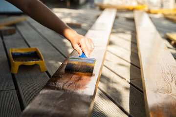 Close-up of hand applying protective wood stain with brush on wooden planks outdoors, DIY renovation in summer
