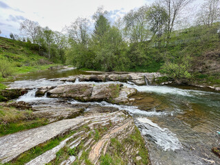 Picturesque stream flows over flat, layered rocks, forming gentle cascades in lush, green landscape. Trees and shrubs adding to natural beauty of scene. Clear water reflects serene surroundings.
