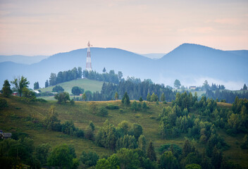 Peaceful mountain landscape at dawn, with rolling hills covered in lush greenery. Communication tower stands atop hill, surrounded by dense trees. Distant mountains enveloped in soft, blue haze.