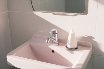 Fototapeta premium Minimal bathroom interior with a pastel pink sink, chrome faucet and white soap dispenser bathed in soft natural light creating gentle shadows and serene minimalist atmosphere.