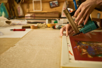 Man using pneumatic stapler to secure canvas print on wooden frame, hands visible working carefully in art studio, focusing on precise assembly process