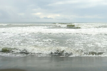 Majestic ocean foamy waves rhythmically crash against a dark black sandy beach in overcast weather. Amazing natural seascape in stormy day. Wild untouched volcanic beach. Cloudy sky under sea.