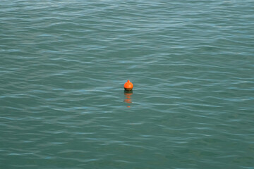 Orange buoy floating on calm water surface with small ripples. Orange maritime safety buoy gently swims on slightly dark turquoise sea, providing navigational guidance across serene oceanic expanse.
