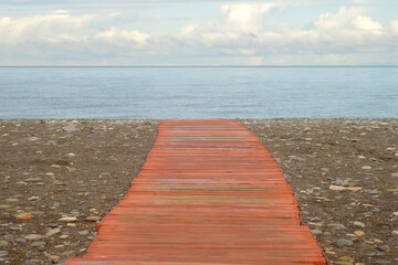Red wooden boardwalk leading to the blue sea under white gray cumulus cloudy sky. Pebble ocean beach with serene and inviting coastal scene. Empty sea shore in overcast weather.