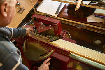 Senior man operating professional framing machine, aligning decorative wooden frame for precise cutting, working with hands on equipment in workshop environment