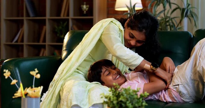 Indian mother tickling son while sitting in modern home on sofa, bonding in quality time together, sharing laughter, affection, love, joy, warmth, parent child relationship, family lifestyle indoors