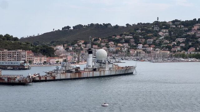 Lateral tracking shot from a boat passing by decommissioned, rusty Cold War-era naval warships in the ship graveyard of Toulon Bay, France.