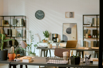 Modern office workspace featuring two desks with laptops, coffee cups, potted plants, stationery, bookshelves, wall clock, copy space on wall, no people visible