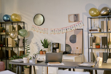 Modern office workspace decorated for birthday celebration with colorful balloons, festive banner reading happy birthday, potted plants, shelves with folders and supplies visible