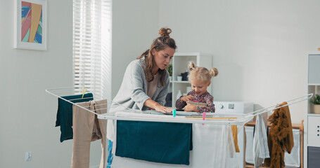 A loving mom dries laundry with the help of her daughter in the bathroom. The girl holds a basket...