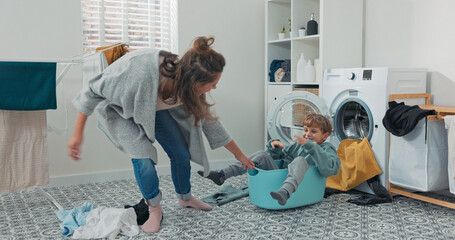 A smiling boy sits in a laundry bowl while his mother pulls him across the bathroom floor. They...