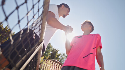 Two athletes train on a sunlit tennis court. A coach in a white shirt and cap guides a player in a bright pink shirt, focusing on skill and teamwork, captured from below.