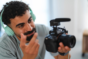 A focused young man wearing headphones holds a camera with a microphone, actively filming or recording content. He gestures while concentrating on his work.