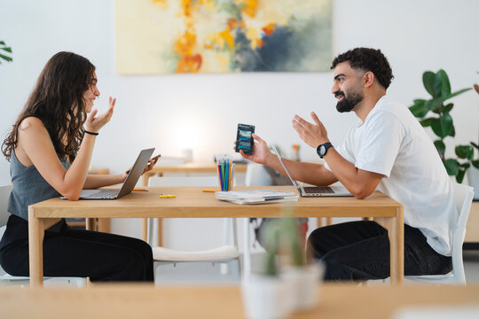 Two young colleagues are having a lively discussion at a modern office table. The man shows something on his phone while the woman listens attentively, both gesturing.