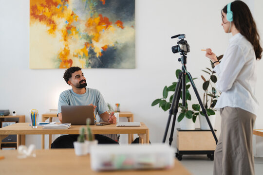 Two people collaborate on a video project. A man works on his laptop while a woman with headphones records content using a camera on a tripod.