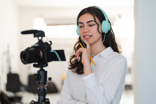 A young woman in a white shirt and light blue headphones holds a pencil, looking focused. She is recording a video or podcast with a camera on a tripod in the background.
