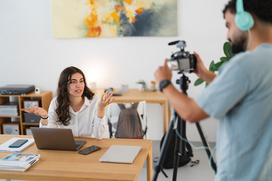 A young woman records a video or podcast at her desk, speaking and gesturing. A man films her with a camera on a tripod, wearing headphones. They are creating content.