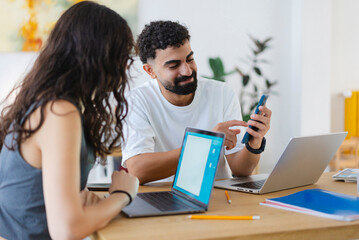 Two young professionals collaborate at a modern office desk. The man smiles while showing content on his smartphone to his female colleague, who is also working on her laptop.