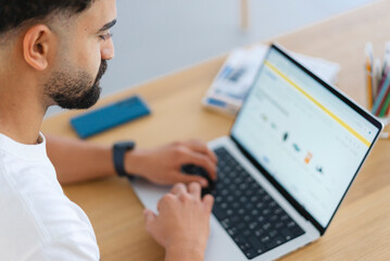 A focused young man with a beard types on his laptop at a wooden desk. He is browsing products online, possibly shopping or working. His smartphone rests nearby.