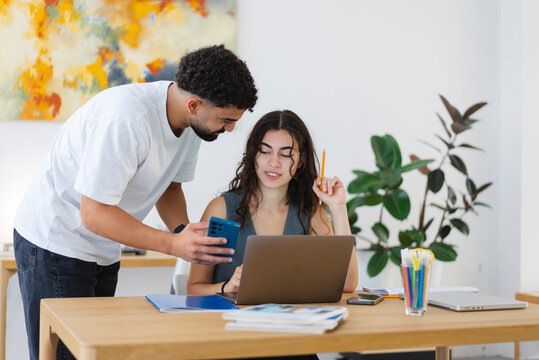 A young man shows his smartphone to a young woman working on a laptop at a desk. They are collaborating in a bright office space with an abstract painting.