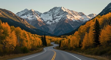 Fototapeta premium Winding road through autumn forest leading to snow capped mountains under clear sky