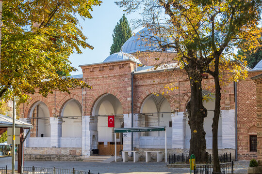 Exterior view of Hamza Bey Mosque in Bursa with its distinctive brickwork, Turkish flag, and surrounding trees.