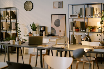 Modern office workspace featuring empty desks with laptops, documents, and stationery, surrounded by shelves filled with folders, potted plants, and minimalist wall art