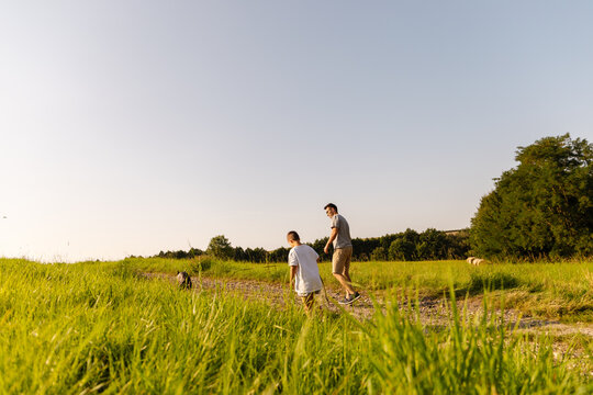 A man and his young son stroll through a lush green field with their dog. The sun shines brightly, creating a warm atmosphere as they spend quality time together outdoors