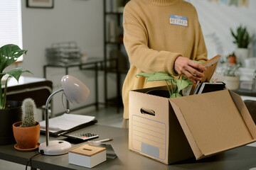 Middle aged Caucasian woman packing personal belongings into cardboard box on office desk, standing near potted plants and desk lamp, wearing badge labeled retired