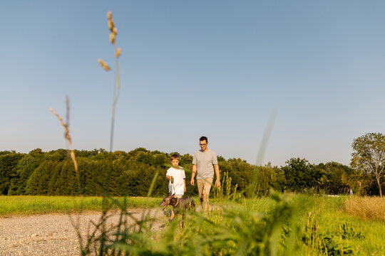 A man and his young son stroll together along a gravel path, holding the leash of their dog. Tall grass sways gently in the breeze under the clear blue sky
