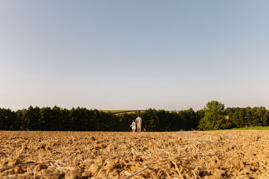 A man and his young son walk together with their dog through a dry field under a clear blue sky. The surrounding trees create a peaceful rural backdrop - Powered by Adobe