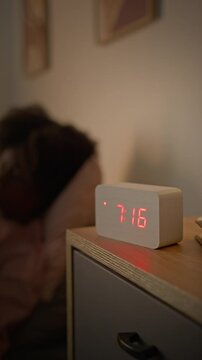 Medium vertical shot of sleepy anonymous young black woman waking up in bed in morning, getting up and fumbling around to switch off alarm clock