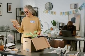 Senior Caucasian woman wearing glasses standing at desk holding framed photo while packing belongings into cardboard box, Black man working on laptop in background, retirement banner visible
