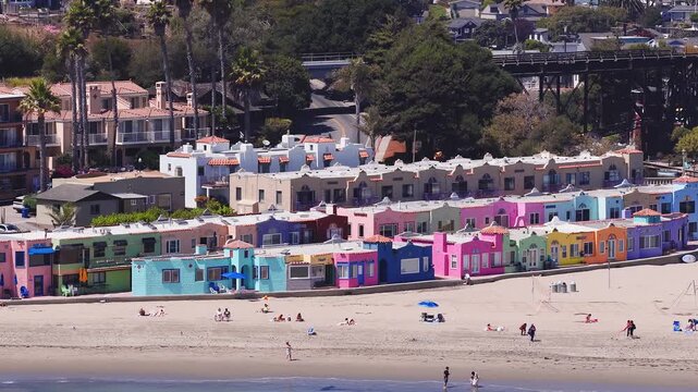 Vibrant pastel buildings, a wooden trestle bridge, sandy beach, and calm river in sunny Capitola, California, with people enjoying outdoor activities.