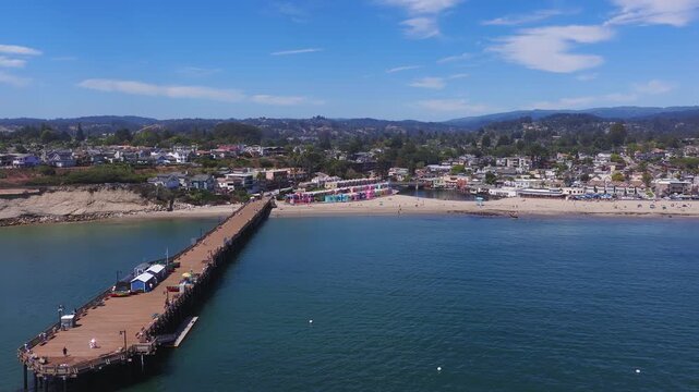 Aerial view of Capitola, California on a sunny day, showing colorful beachfront buildings, a sandy beach, a wooden pier, and calm blue ocean.