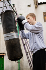 Young man in boxing gloves intensely training with a worn punching bag outdoors, showcasing...