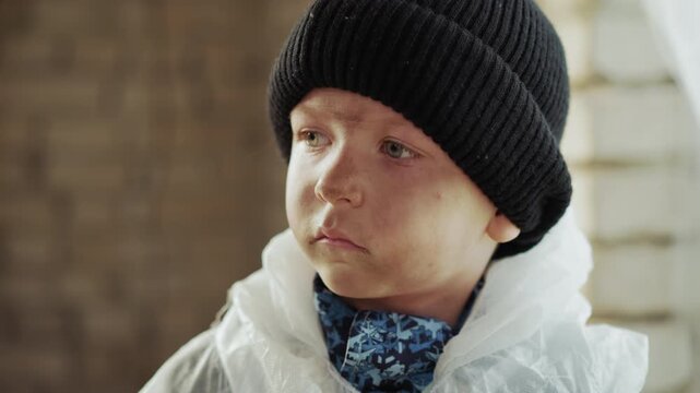 Close up of young boy with dirty face wearing black knitted hat and white protective jacket, staring ahead with serious focused expression against blurred background