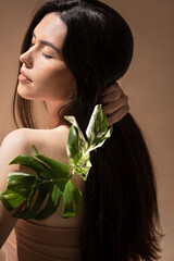 Woman with long dark hair gently touching her neck, adorned with a green leaf, against a soft beige background, embodying natural beauty and tranquility in a serene moment