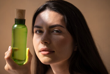 Young woman with long dark hair holds a green bottle with wooden cap, showcasing natural beauty product in soft light, emphasizing skincare and wellness concept