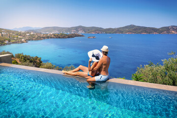 A hugging couple sits on a swimming pool edge and looks over the sea during their summer holidays