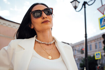 Confident woman wearing sunglasses and layered necklaces poses stylishly outdoors, showcasing modern fashion and urban lifestyle against a vibrant city backdrop