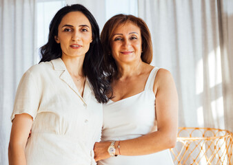 Two women, one in a light dress and the other in a casual outfit, stand together in a bright, airy room, showcasing warmth and connection in a modern interior setting
