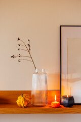 Decorative shelf arrangement featuring a clear glass vase with branches, a yellow gourd, a small orange pumpkin, and a lit candle creating a cozy autumn atmosphere