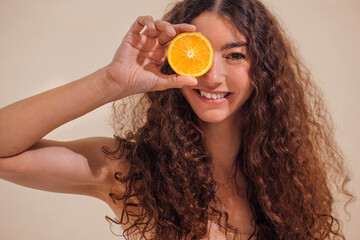 Young woman with curly hair playfully holding a slice of orange in front of her face, smiling brightly, showcasing a vibrant and cheerful atmosphere with fresh fruit