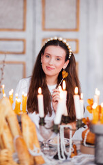Young woman with long hair, wearing a pearl headband, is smiling while holding a heart-shaped lollipop, surrounded by candles and decorative pastries, creating a warm atmosphere