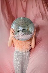 Young girl wearing a ruffled top and sparkling pants holds a large disco ball in front of her face, surrounded by soft pink fabric, creating a whimsical and playful atmosphere