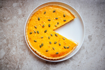 Delicious pumpkin pie with a slice removed, garnished with pumpkin seeds, displayed on a white plate against a textured gray background, showcasing culinary artistry and dessert appeal