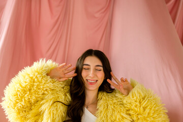Young woman with long dark hair wearing a fluffy yellow coat is playfully posing against a soft pink backdrop, showcasing a joyful and vibrant atmosphere of self-expression
