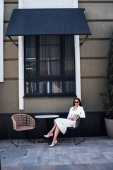 Stylish Asian woman in white dress and sunglasses sitting at a table outside a modern building, enjoying a sunny day, exuding confidence and elegance in urban setting